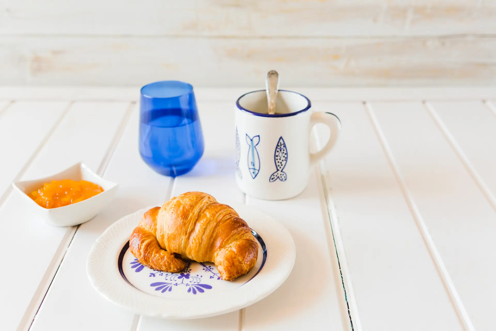 La colazione sul tavolo bianco: cornetto nel piatto dipinto di blu, tazza col pesce disegnato, bicchiere blu e marmellata